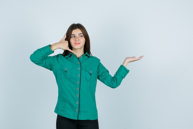 Jovem garota na blusa verde, calça preta, mostrando o gesto do telefone, espalhando a palma da mão para o lado e parecendo otimista, vista frontal.