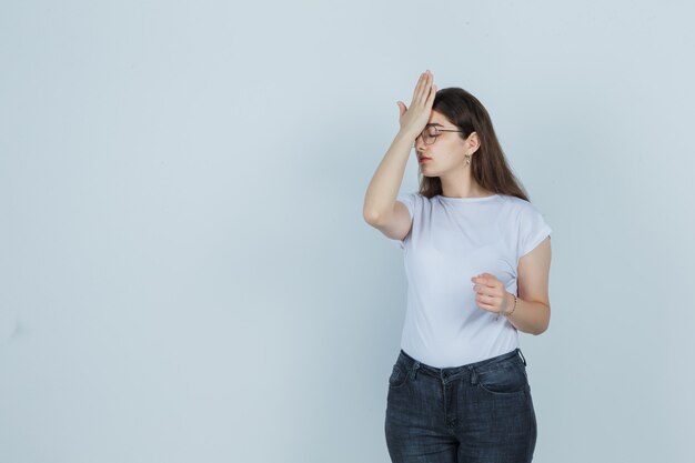 Jovem garota segurando a mão na testa em t-shirt, jeans e parecendo cansada. vista frontal.