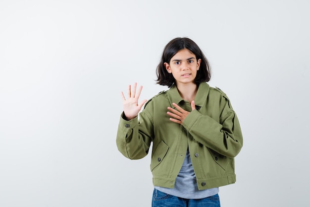 Jovem garota segurando a mão no peito e fazendo promessa ou juramento em suéter cinza, jaqueta cáqui, calça jeans e olhando séria, vista frontal.