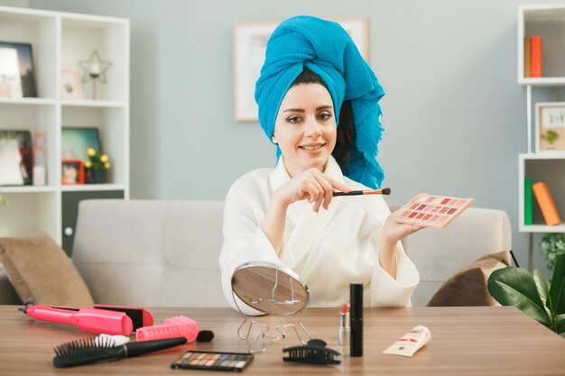 Jovem garota segurando paleta de sombra com pincel de maquiagem enrolado em uma toalha no cabelo, sentada à mesa com ferramentas de maquiagem na sala de estar