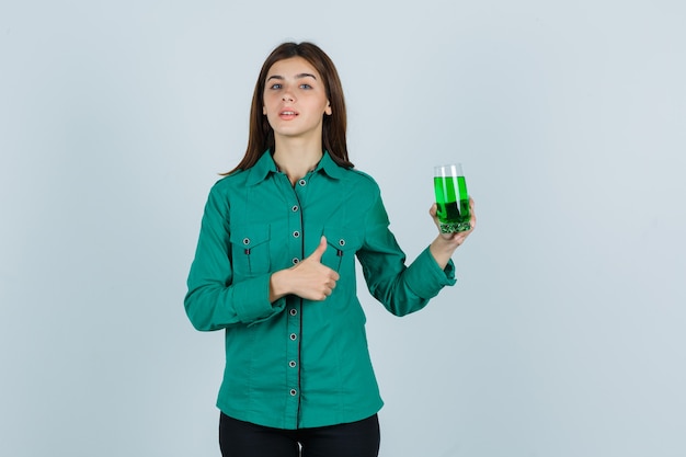 Foto grátis jovem garota segurando um copo de líquido verde, aparecendo o polegar na blusa verde, calça preta e olhando sério. vista frontal.