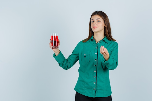 Jovem garota segurando um copo de líquido vermelho, mostrando gesto italiano na blusa verde, calça preta e parecendo satisfeita. vista frontal.