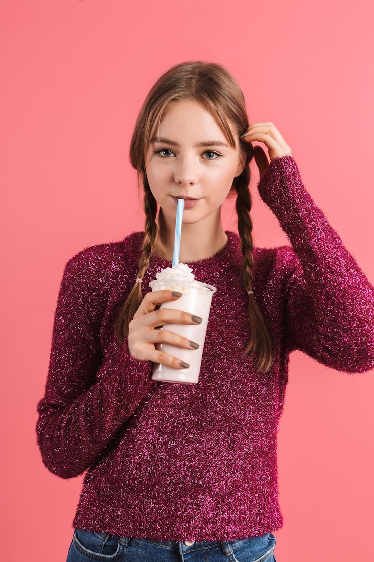 Foto grátis jovem garota sorridente atraente com duas tranças na camisola segurando milk-shake nas mãos enquanto sonhadoramente olhando na câmera sobre fundo rosa