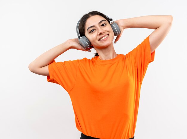 Jovem garota vestindo uma camiseta laranja com fones de ouvido feliz e positiva curtindo sua música favorita sorrindo em pé sobre uma parede branca