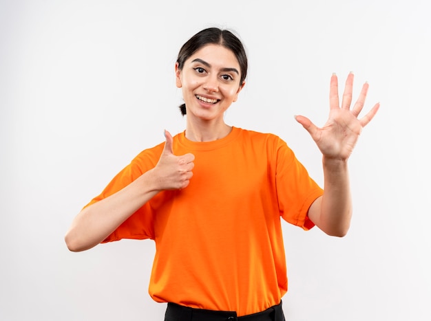 Jovem garota vestindo uma camiseta laranja olhando para a câmera e sorrindo, mostrando o número seis em pé sobre um fundo branco