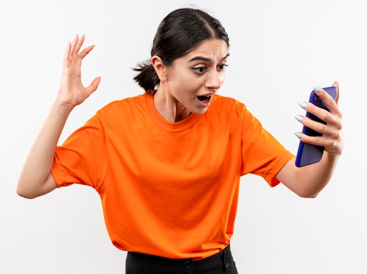 Foto grátis jovem garota vestindo uma camiseta laranja olhando para a tela do smartphone animada e confusa com a mão levantada em pé sobre uma parede branca