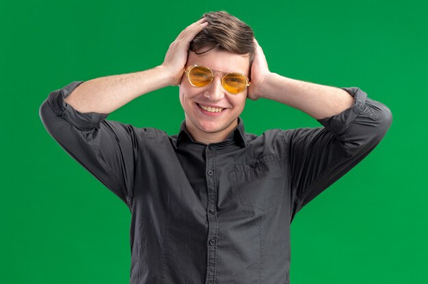 Foto grátis jovem garoto caucasiano sorridente usando óculos de sol, colocando as mãos na cabeça e olhando para a câmera