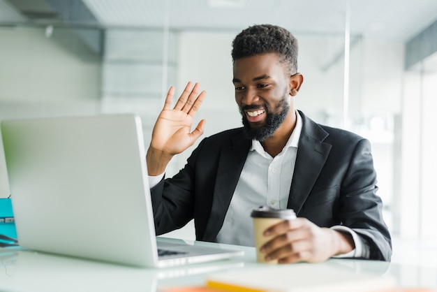 Jovem gerente afro-americano com restolho sentado na frente do laptop aberto usando fones de ouvido enquanto faz uma videoconferência com parceiros de negócios