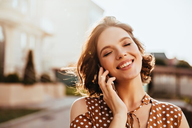 Jovem glamorosa falando no telefone com os olhos fechados. Foto ao ar livre de uma garota bonita caucasiana com cabelo castanho curto.