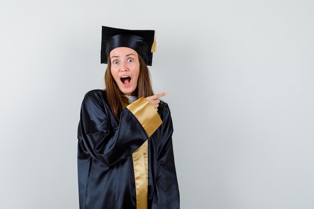 Jovem graduada feminina apontando para o canto superior direito em traje acadêmico e parecendo surpresa. vista frontal.