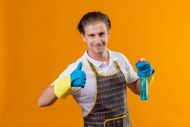 Jovem hansdome usando avental e luvas de borracha segurando spray de limpeza, feliz e sorrindo positivamente mostrando polegares para cima em pé sobre a parede laranja
