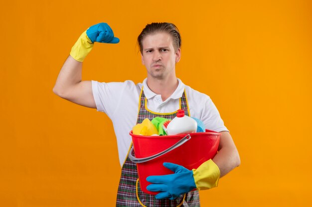 Jovem hansdome usando avental e luvas de borracha, segurando um balde com ferramentas de limpeza, levantando o punho com uma expressão séria