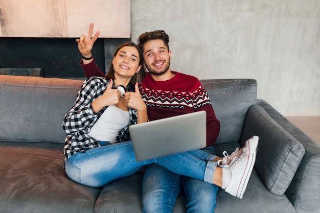 Jovem hippie sorrindo, homem e mulher, sentado em casa no inverno, segurando um laptop