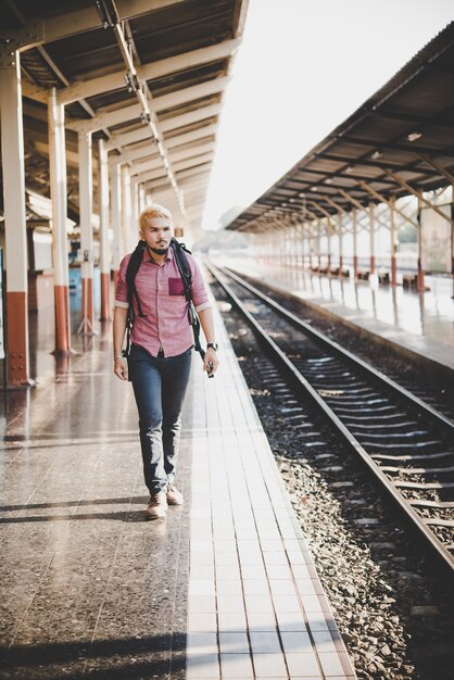 Jovem hipster que espera na plataforma da estação com mochila. Conceito de viagem.