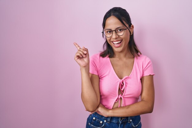 Jovem hispânica em pé sobre fundo rosa usando óculos com um grande sorriso no rosto, apontando com a mão e o dedo para o lado olhando para a câmera.