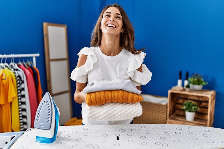 Foto grátis jovem hispânica passando roupas na lavanderia segurando suéteres dobrados sorrindo e rindo muito alto porque uma piada maluca