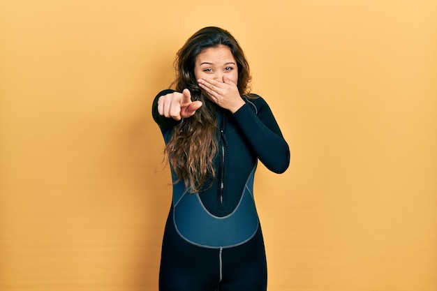 Jovem hispânica vestindo uniforme de neoprene mergulhador rindo de você, apontando o dedo para a câmera com a mão sobre a boca, expressão de vergonha