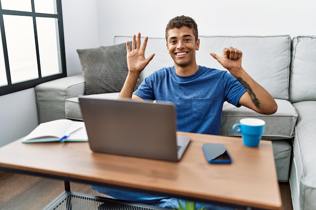 Jovem hispânico bonito usando laptop sentado no chão mostrando e apontando para cima com os dedos número seis enquanto sorria confiante e feliz.