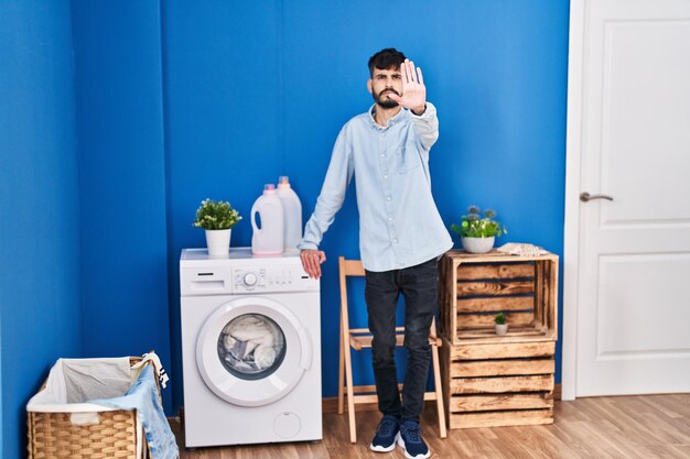 Jovem hispânico com barba lavando roupa em pé na lavanderia com a mão aberta fazendo sinal de pare com gesto de defesa de expressão séria e confiante