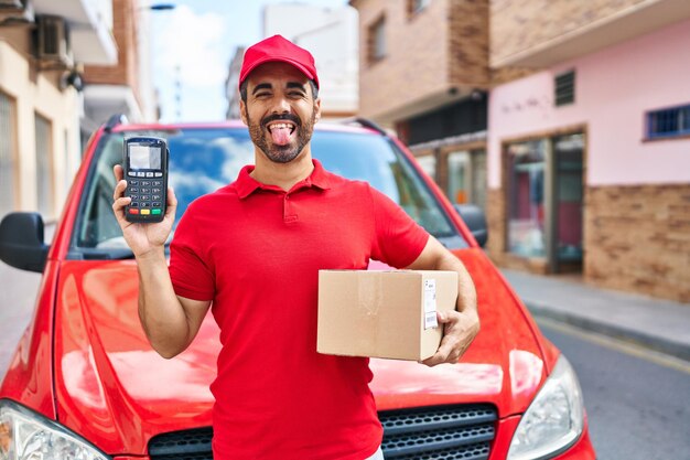 Jovem hispânico com barba vestindo uniforme de entrega e boné segurando telefone de dados enfiando a língua feliz com expressão engraçada