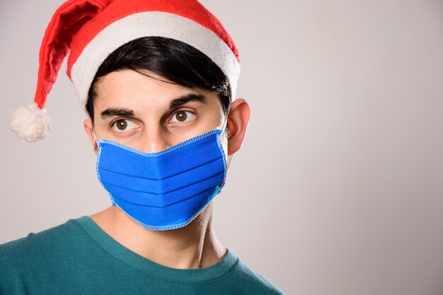 Jovem hispânico com máscara facial e chapéu de papai noel olhando para o lado