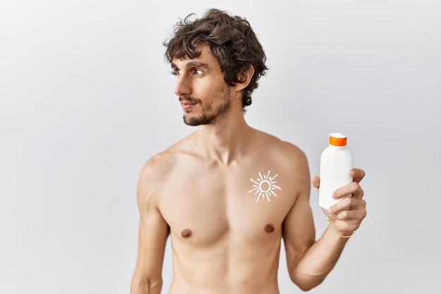 Jovem hispânico em pé sem camisa segurando loção protetor solar olhando para o lado, relaxe a pose de perfil com rosto natural e sorriso confiante.