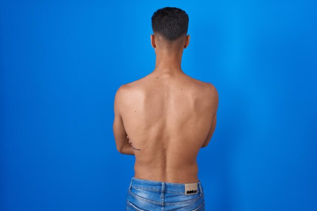 Jovem hispânico em pé sem camisa sobre fundo azul em pé para trás olhando para longe com os braços cruzados