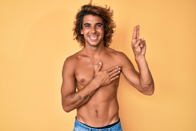 Jovem hispânico em pé sem camisa sorrindo xingando com a mão no peito e os dedos para cima, fazendo um juramento de promessa de lealdade
