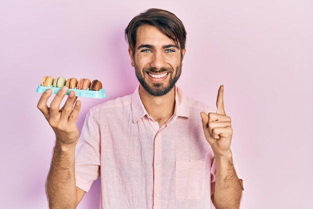 Jovem hispânico segurando deliciosos bolos macrons sorrindo com uma ideia ou pergunta apontando o dedo com cara feliz, número um