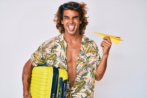 Jovem hispânico segurando mala e avião de papel saindo de férias de verão mostrando a língua feliz com expressão engraçada