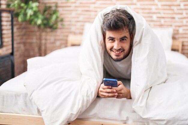 Jovem hispânico usando smartphone deitado na cama no quarto