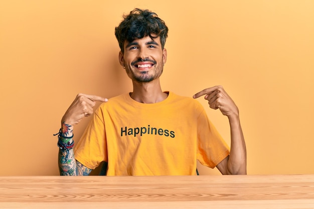 Jovem hispânico vestindo camiseta com mensagem de felicidade sentado na mesa parecendo confiante com sorriso no rosto apontando-se com os dedos orgulhoso e feliz