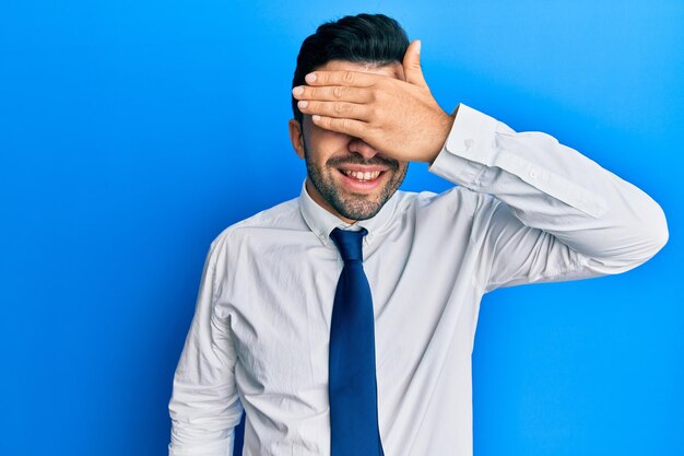 Jovem hispânico vestindo roupas de negócios sorrindo e rindo com a mão no rosto cobrindo os olhos para o conceito cego de surpresa