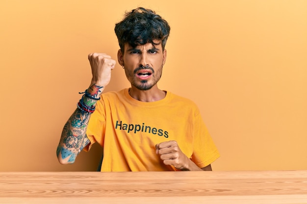 Jovem hispânico vestindo t-shirt com mensagem de palavra de felicidade sentado na mesa zangado e louco levantando o punho frustrado e furioso enquanto gritava com raiva raiva e conceito agressivo
