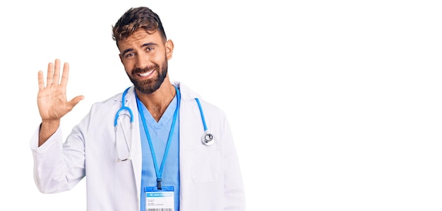 Jovem hispânico vestindo uniforme médico e estetoscópio mostrando e apontando para cima com os dedos número cinco enquanto sorria confiante e feliz.