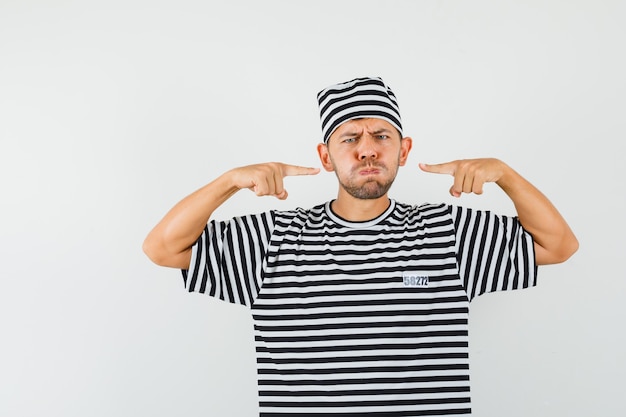 Jovem homem apontando para as bochechas dilatadas com um chapéu de camiseta listrada e parecendo melancólico