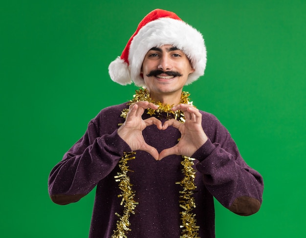 Jovem homem bigodudo usando chapéu de Papai Noel de natal com enfeites em volta do pescoço olhando para a câmera com uma cara feliz sorrindo fazendo gesto de coração com os dedos em pé sobre um fundo verde