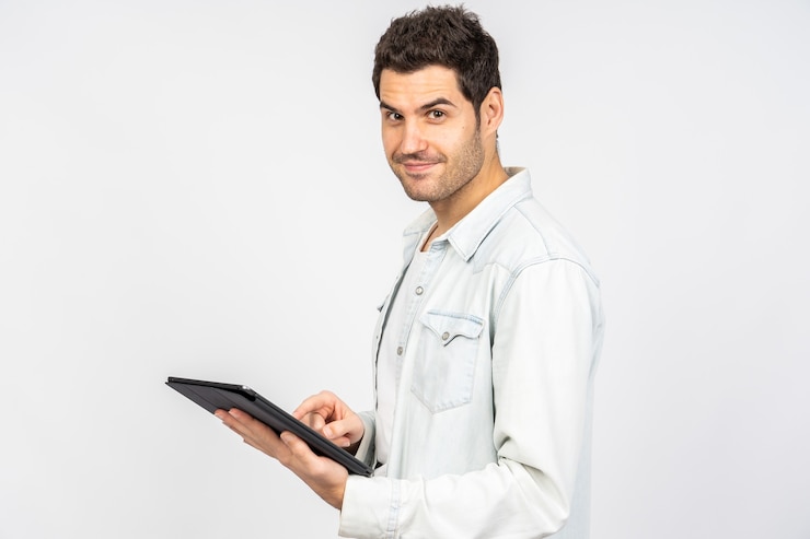 Foto grátis jovem homem branco sorrindo enquanto trabalha em um tablet contra uma parede branca