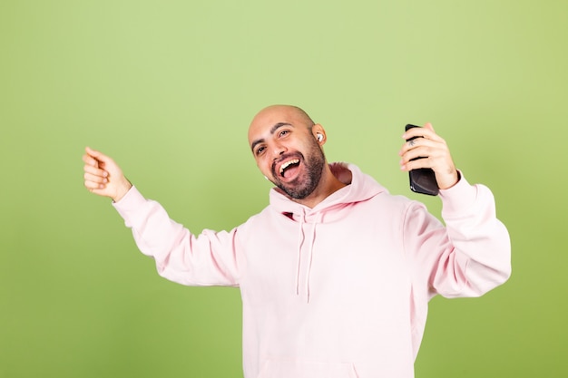 Jovem homem careca, caucasiano, com capuz rosa isolado, segurando o telefone, feliz, dançando, movendo-se em fones de ouvido, curtindo com os olhos fechados