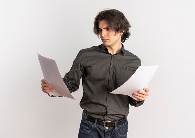 Jovem homem caucasiano bonito irritado segurando e olhando para folhas de papel em branco isoladas no fundo branco com espaço de cópia