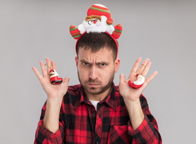 Jovem homem caucasiano com a testa franzida usando uma bandana de papai noel segurando enfeites de natal de papai noel na parede branca