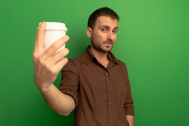 Jovem homem caucasiano satisfeito esticando a xícara de café de plástico em direção à câmera, olhando para a câmera isolada em um fundo verde com espaço de cópia