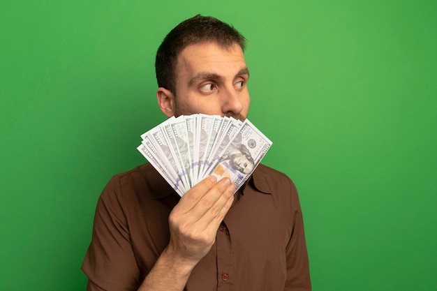Jovem homem caucasiano segurando dinheiro olhando para o lado por trás, isolado em um fundo verde com espaço de cópia