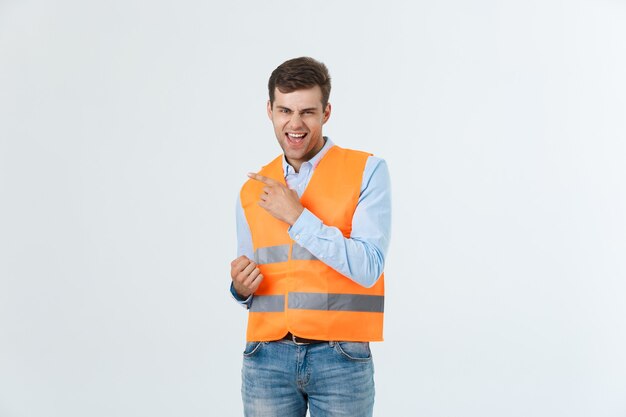 Jovem homem caucasiano sobre fundo branco, vestindo uniforme de contratante e capacete de segurança, surpreso com uma ideia ou pergunta, apontando o dedo com uma cara feliz, número um.