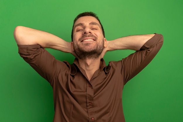 Foto grátis jovem homem caucasiano sorridente, olhando para a câmera, colocando as mãos atrás da cabeça isoladas sobre fundo verde