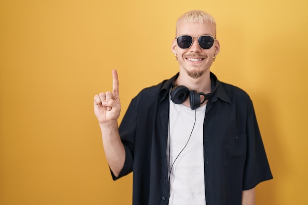 Jovem homem caucasiano usando óculos escuros em pé sobre fundo amarelo mostrando e apontando para cima com o dedo número um enquanto sorria confiante e feliz.