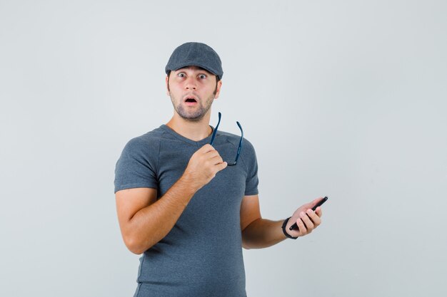 Jovem homem com boné de camiseta cinza segurando um celular e óculos e parecendo surpreso