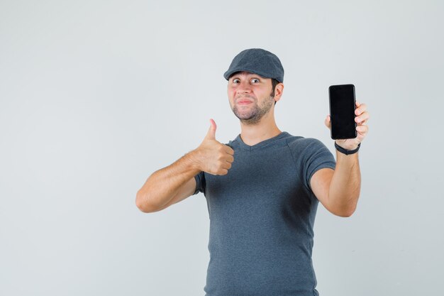 Jovem homem com boné de camiseta mostrando o polegar enquanto segura um telefone celular