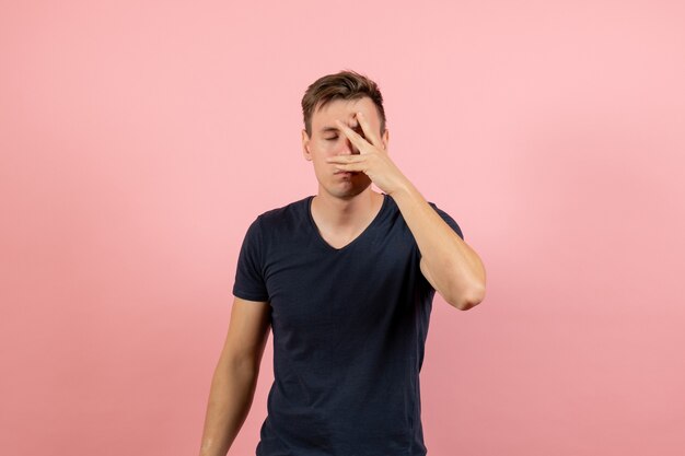 Jovem homem com camisa azul escura, vista frontal, cobrindo o rosto no fundo rosa, modelo masculino de cor de emoção humana