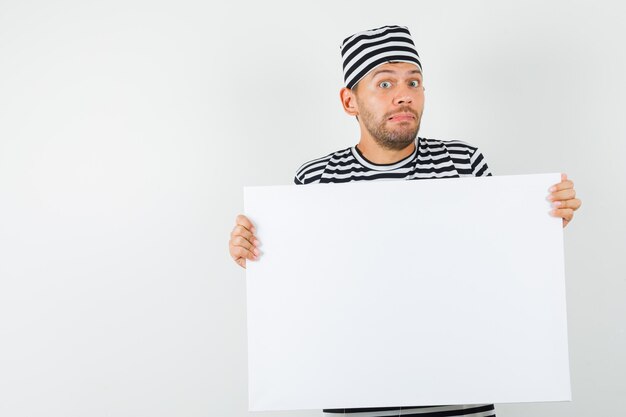 Jovem homem com chapéu de camiseta listrada segurando uma tela em branco e parecendo confuso
